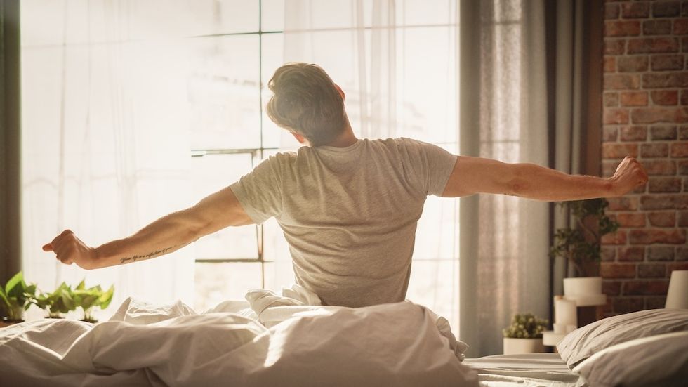 A young man waking up in the morning with the sun shining through his window, sitting up in bed stretching.