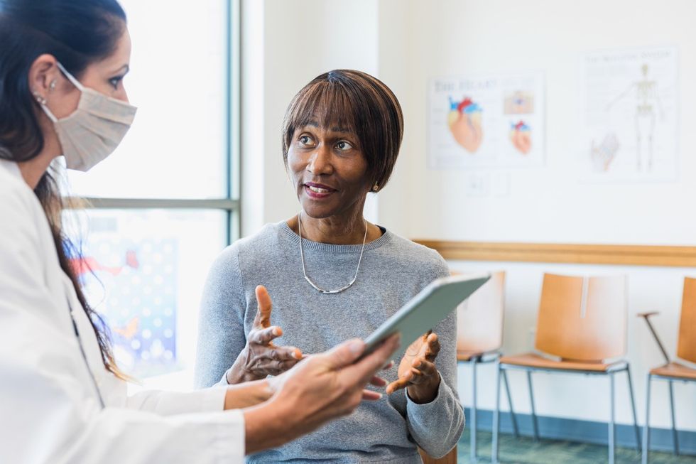 An attentive doctor listens as a female patients asks questions about recent medical test results. The doctor is holding a digital tablet which contains the test results. The doctor is wearing a protective face mask.