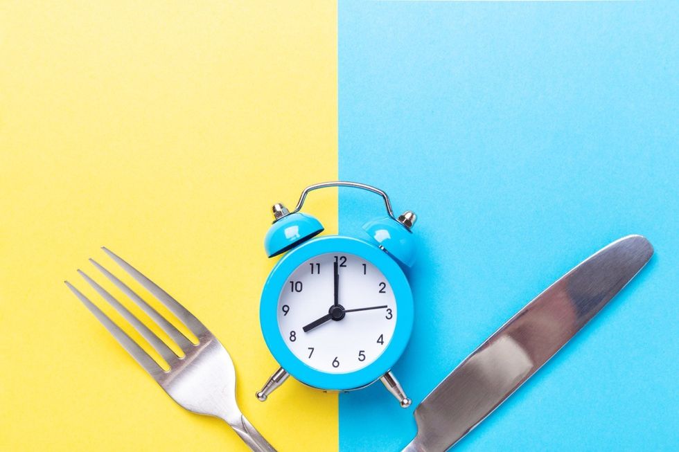 Blue alarm clock, fork, and knife on yellow and blue background. Intermittent fasting concept.