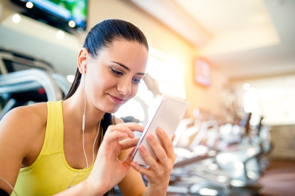 woman in a yellow tank sitting on the edge of a treadmill at the gym using her phone