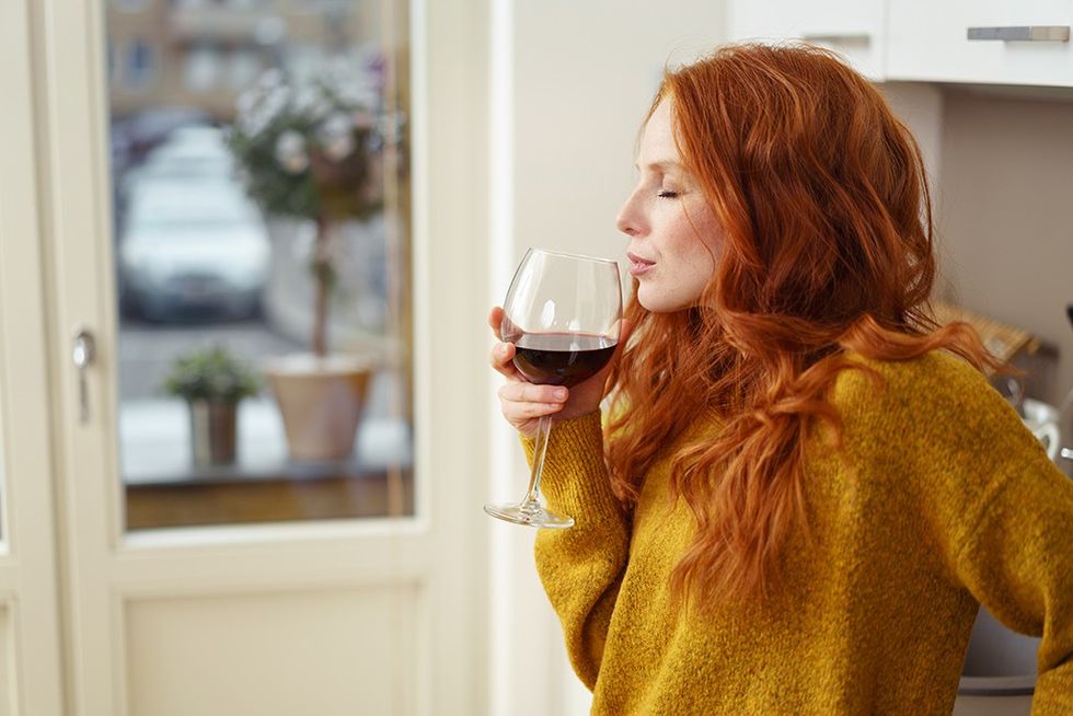 woman with red hair wearing a mustard-color sweater sipping a glass of red wine