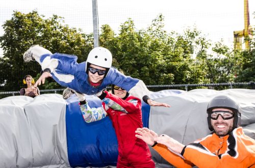 A woman, dressed like a skydiver, is feeling the free falling with the free falling simulator (huge fan) and with the help of two persons responsible of the simultator. This is happening outside during a summer day.