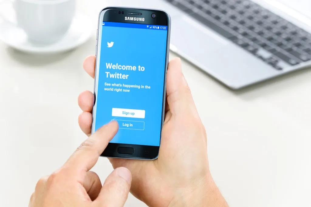 white person's hand holds phone featuring blue Twitter welcome screen, trying to reach a customer service rep
