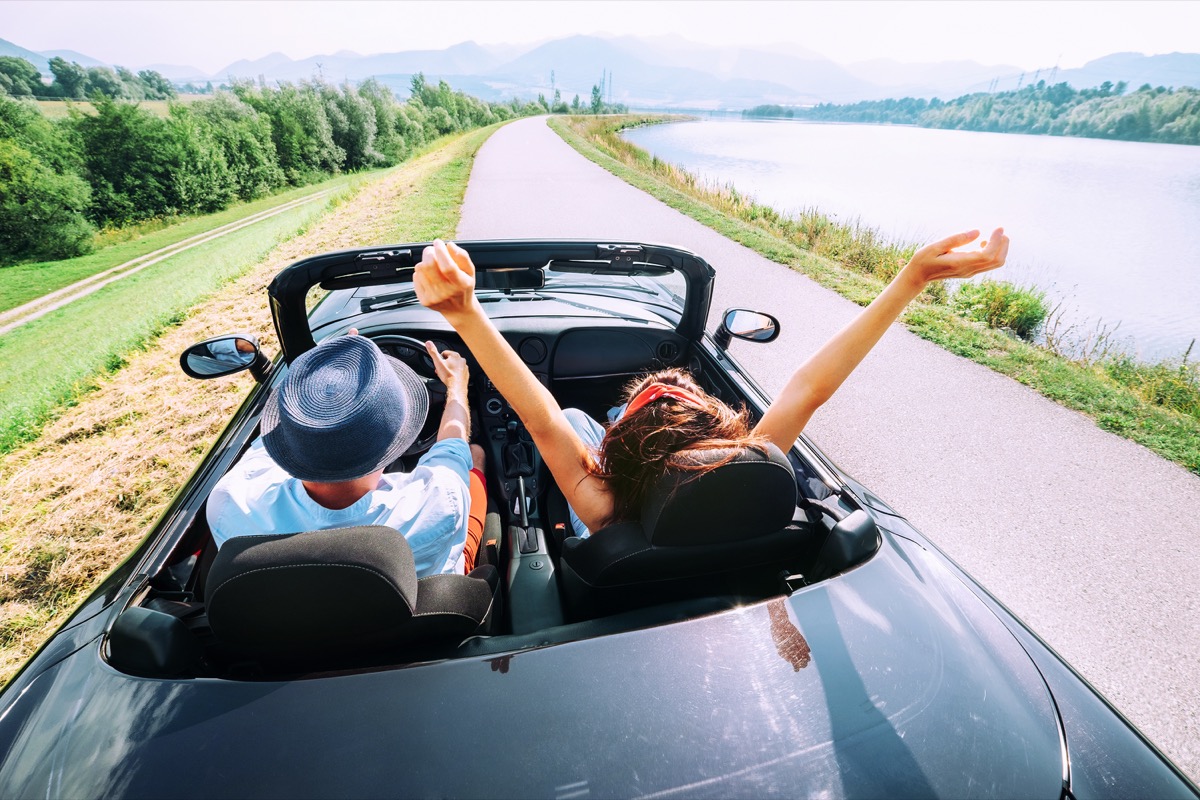 Couple driving a convertible down the road adjacent to the water for a weekend getaway