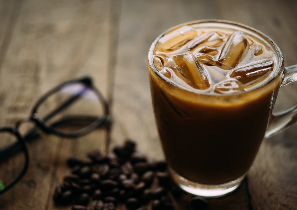 glass mug of iced coffee sitting on wooden table with coffee beans and eyeglasses
