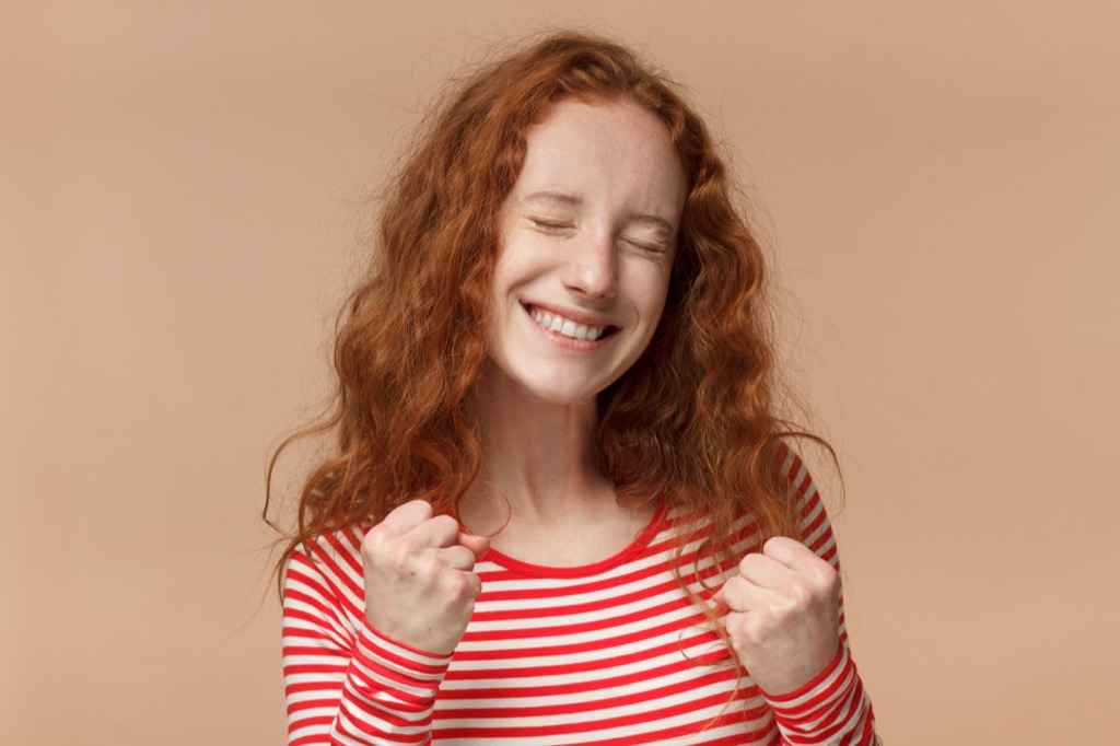 red headed young woman in red and white striped shirt grips fists in excitement as she smiles and closes her eyes