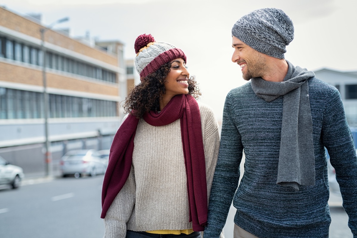 Casual young couple holding hands while walking on the street. Happy couple wearing winter clothes and looking at each other. Romantic multiethnic man and woman enjoying winter breeze outdoor. (Casual young couple holding hands while walking on the st