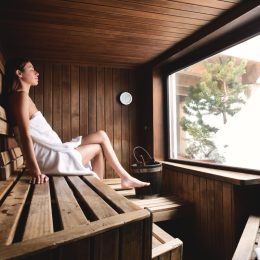 A beautiful woman wearing a white towel takes a sauna: The sauna is made of wood with a large window with a view of the snow.