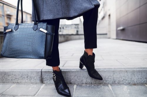 closeup of a woman from her knees down, wearing a long dark gray coat, black pants, black ankle boots, carrying a large black leather purse