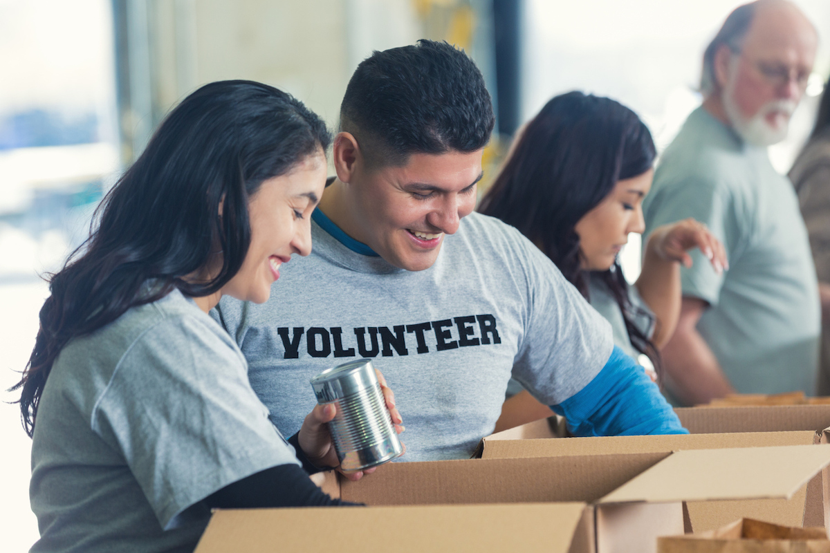 Latinx man and woman are volunteering together in community food bank for charity. Couple is sorting food donations into cardboard boxes, so boxes can be distributed to less fortunate people. They are smiling and enjoying giving back to community. Man is wearing a VOLUNTEER tshirt.
