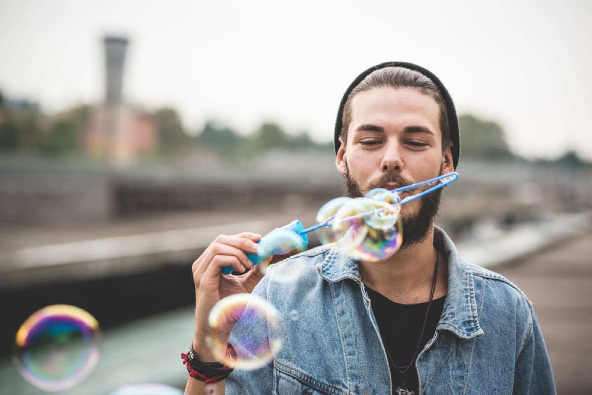 young white hipster wearing beanie and jean jacket blows bubbles in urban environment