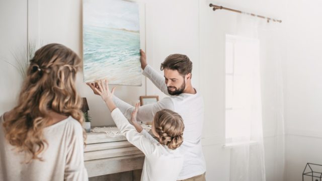 white man, woman, and child hanging painting on white wall over fireplace