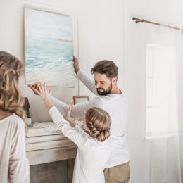 white man, woman, and child hanging painting on white wall over fireplace
