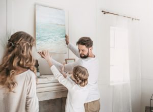 white man, woman, and child hanging painting on white wall over fireplace