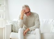 older man sits on a couch and holds his head from stress