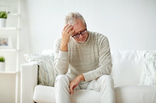 older man sits on a couch and holds his head from stress