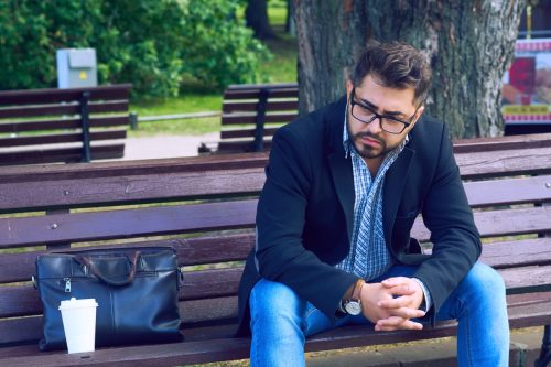 young man in glasses sitting on a bench in the park with sad facial expression. Office worker lost his job. Middle aged man despair economic crisis.