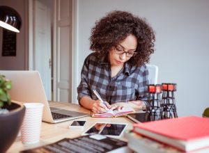 Shot of afro american woman in a home office taking notes in diary. Businesswoman sitting at table and working from home office.