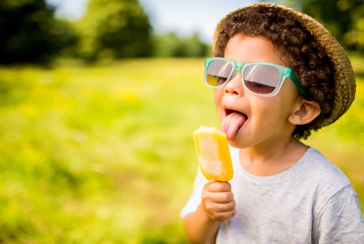 The tongue of a young boy with curly hair is touching an orange frozen treat. He is wearing a gray t-shirt, a straw hat and blue rimmed glasses. The background is blurry and consists of an open field of green grass with trees and bushes in the distance.