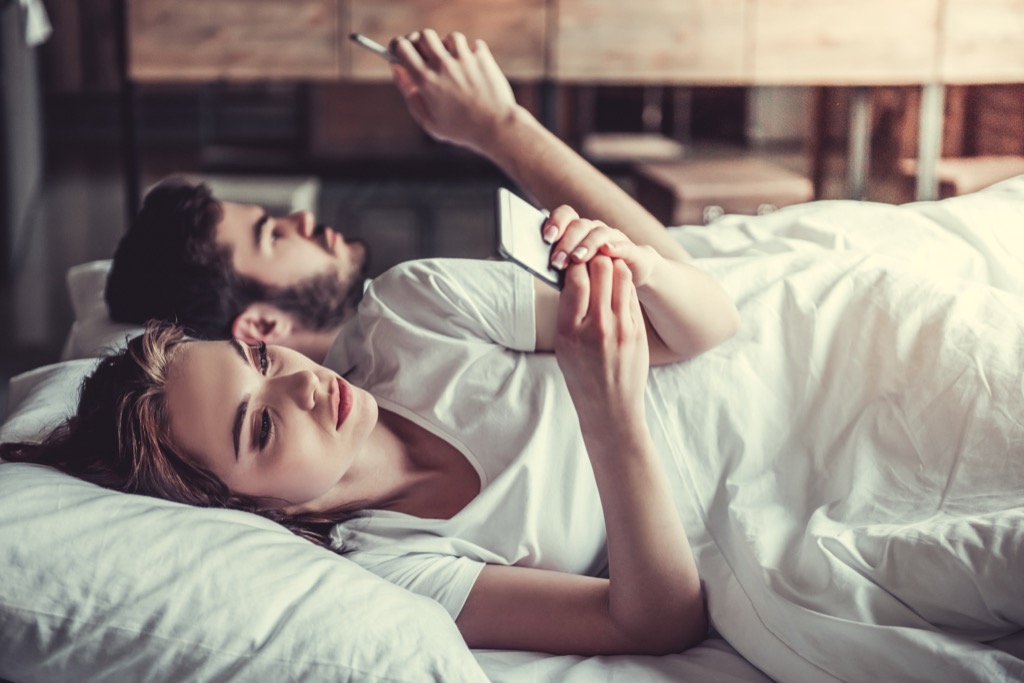 bored couple lying in bed with smartphones, things you should never say to your spouse