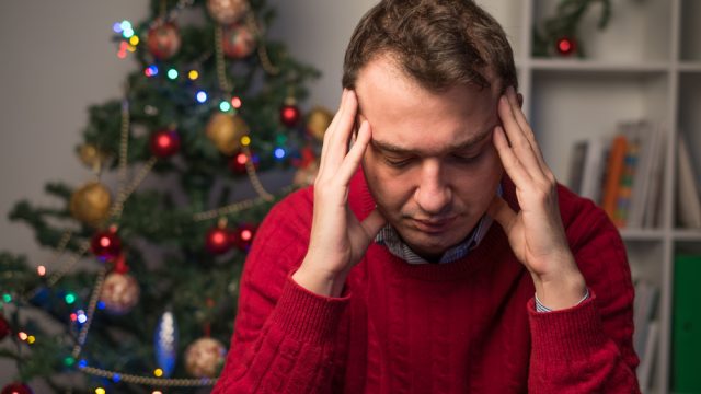 stressed man in red sweater holds head while sitting near christmas tree
