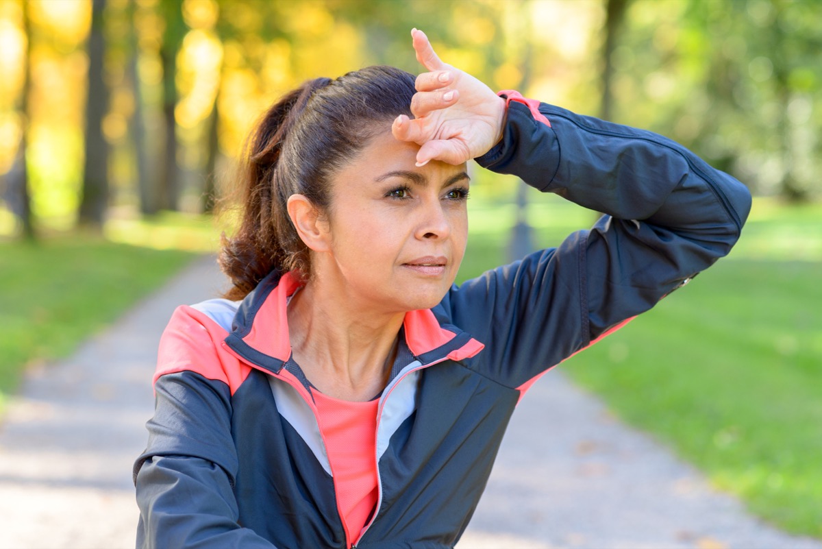 Woman wiping her brow with the back of her hand as she jogs outdoors in a park looking to the side with a thoughtful serious expression