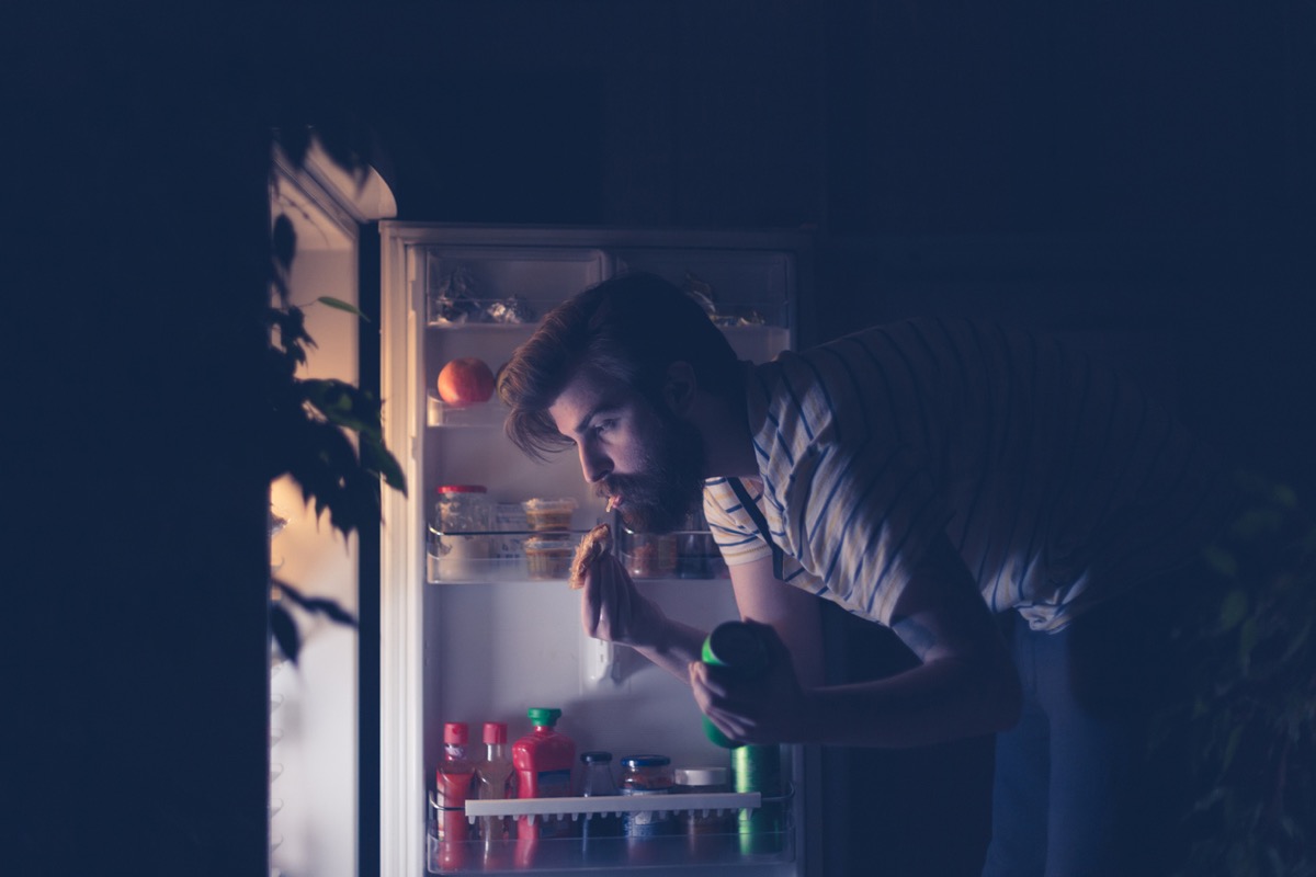 young white man grabbing a snack and a beer from an open fridge in the dark