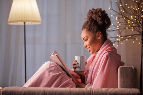 young black woman drinking tea and reading a book in a robe on her couch