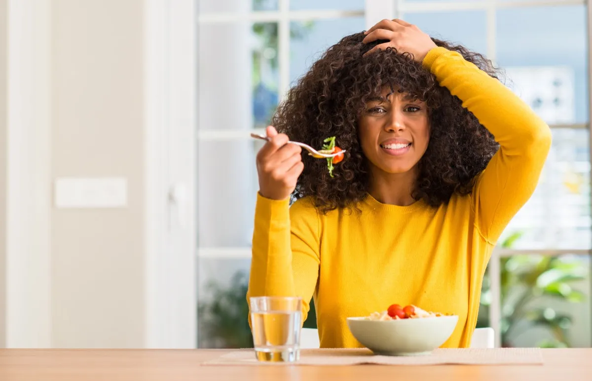 African american woman eating pasta salad at home stressed with hand on head, shocked with shame and surprise face, angry and frustrated. Fear and upset for mistake.