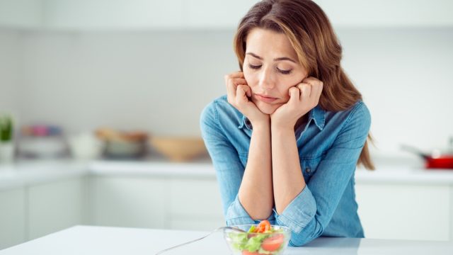 middle aged woman looks disappointed at small salad while standing in kitchen