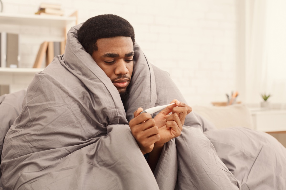 sick black man under a blanket looking at a thermometer