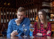 young white man looking at his phone while white girlfriend looks upset at wine bar