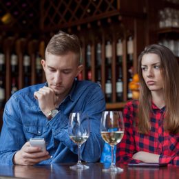 young white man looking at his phone while white girlfriend looks upset at wine bar