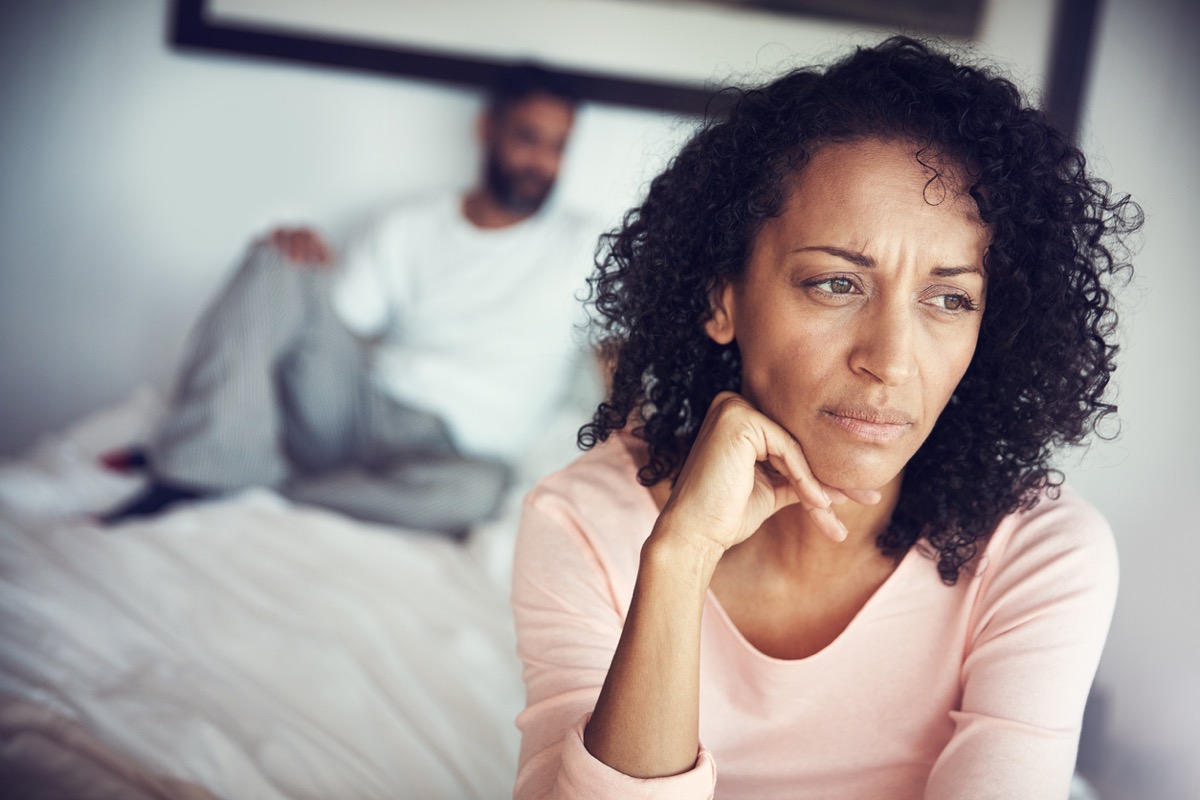 woman looks upset with husband lying on bed behind her