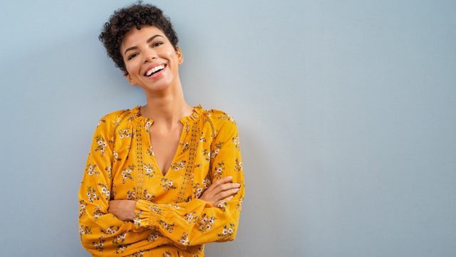 Portrait of beautiful cheerful girl smiling and looking at camera. Happy african woman in casual standing on blue background. Brazilian stylish woman with crossed arms and curly hair isolated with copy space.