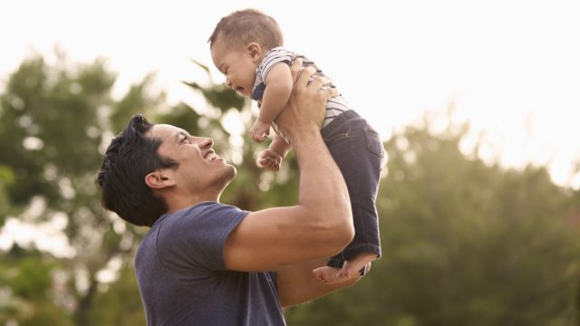 Millennial Hispanic father holding his little baby in the air in the park, close up
