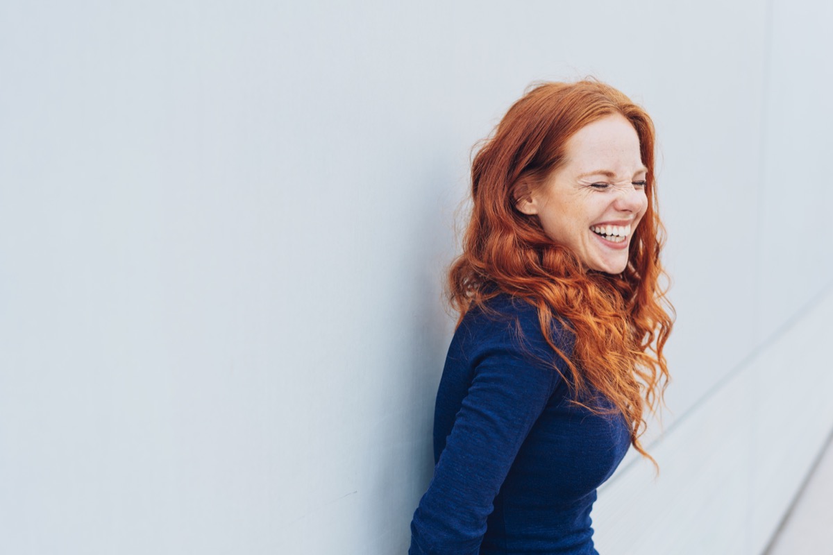 attractive young woman standing giggling or laughing at something she finds very funny while leaning against a white exterior wall with copy space