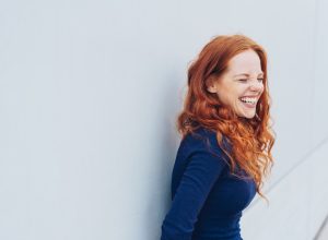 attractive young woman standing giggling or laughing at something she finds very funny while leaning against a white exterior wall with copy space