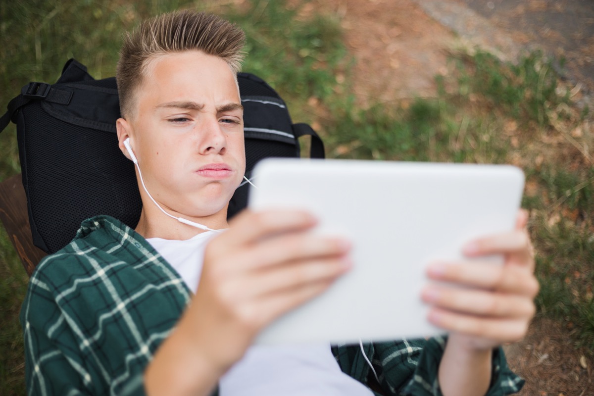 white teen boy looking annoyed at an ipad while lying on his backpack outside