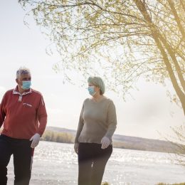 older white man and white woman walking by the water with face masks