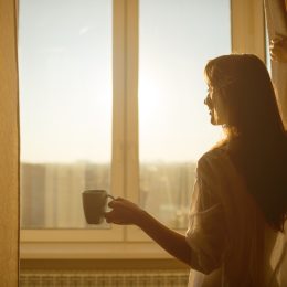 Woman opening curtains in morning with coffee