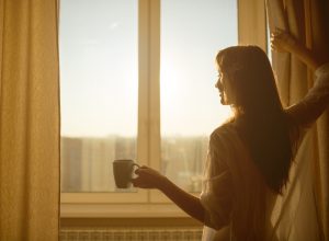 Woman opening curtains in morning with coffee