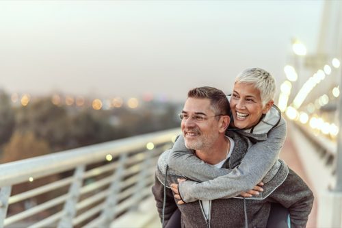 Photo of Happy mature couple having fun in the city on a autumn day. Love story true feelings concept. Portrait of friendly peaceful fitness couple in love. Couple enjoying the outdoors together. Sporty couple in love enjoying each other. Handsome man giving a gray hair woman a piggy back ride.