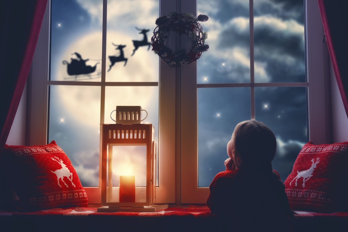 Little girl staring outside her window at Santa's sleigh and reindeer in the sky