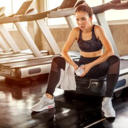 Asian young woman tired taking a break from running or exercise sitting on treadmill machine drinking water and towel sweat in fitness gym healthy .girl in sportswear workout rest in morning