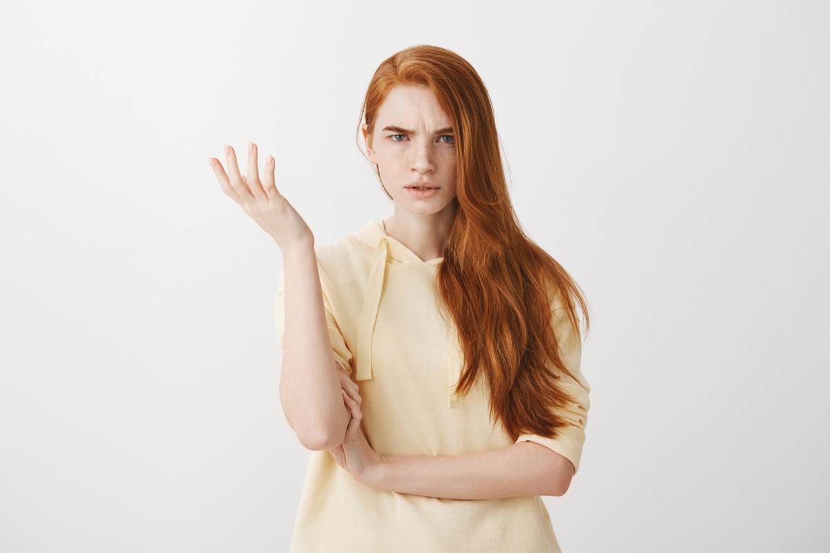 What the heck are you talking about, nonsense. Studio shot of frustrated female with red hair gesturing with raised palm, frowning, being displeased and confused with dumb question over gray wall