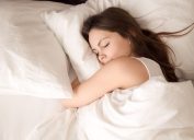 Top view of attractive young woman sleeping well in bed hugging soft white pillow. Teenage girl resting, good night sleep concept. Lady enjoys fresh soft bedding linen and mattress in bedroom