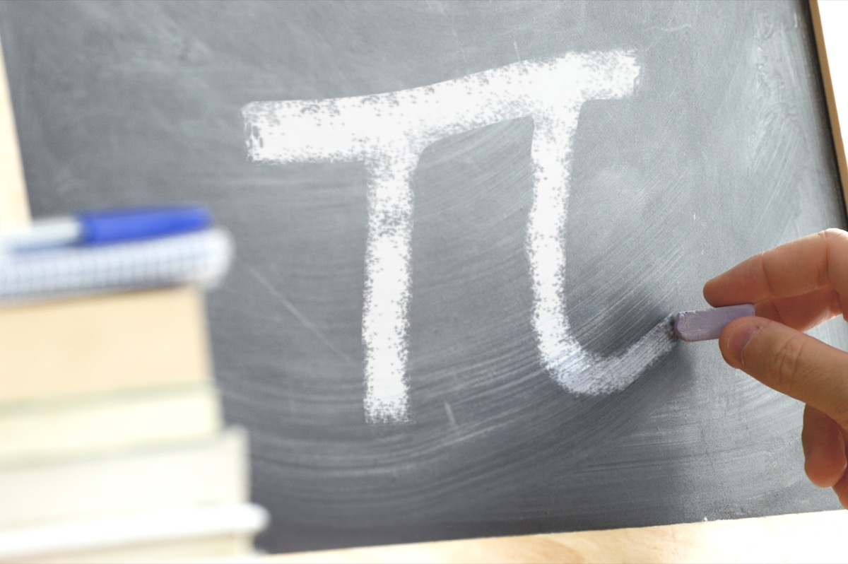 Hand writing on a blackboard in a Math class with the PI symbol written on. Some books and school materials