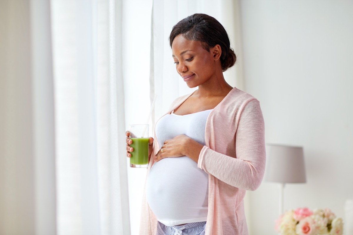 happy pregnant african american woman drinking green vegetable juice or smoothie at home