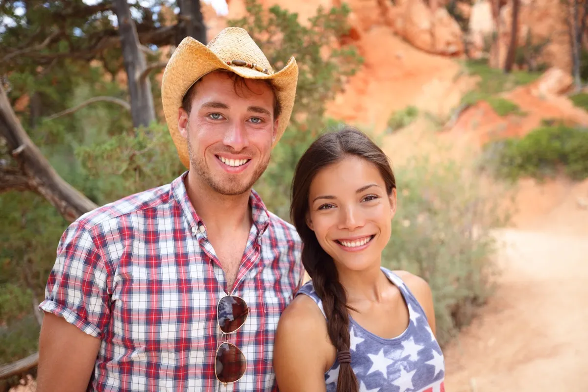Happy outdoors couple portrait in american countryside. Smiling multiracial young couple in western USA nature. Man wearing cowboy hat and woman wearing USA flag shirt